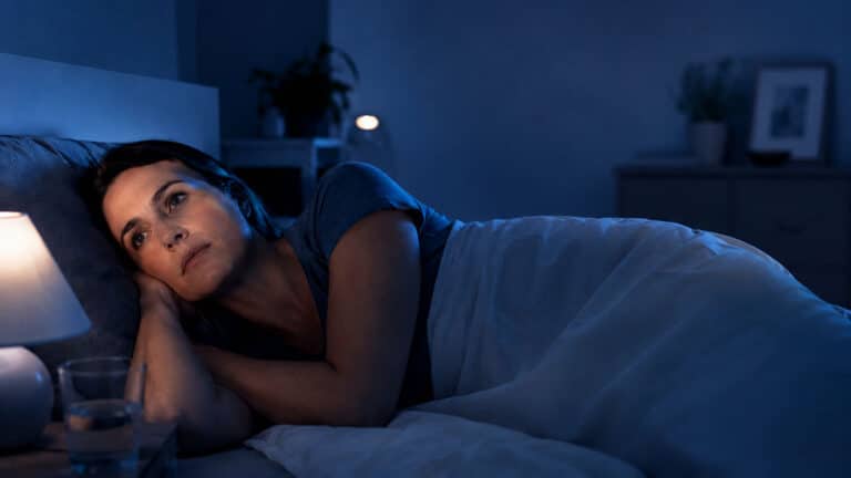 Woman lying awake in bed under blue night lighting, looking worried and downcast.