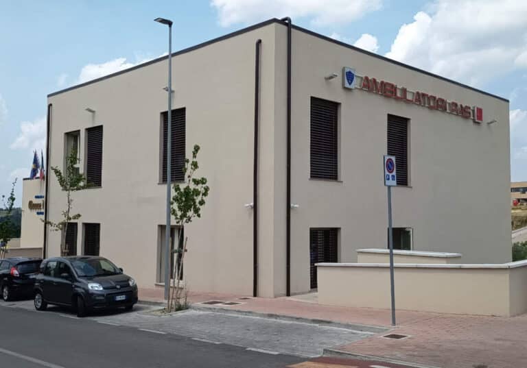 Two-story beige clinic with an 'AMBULATORIO' sign on the corner, parked cars and a street in front.